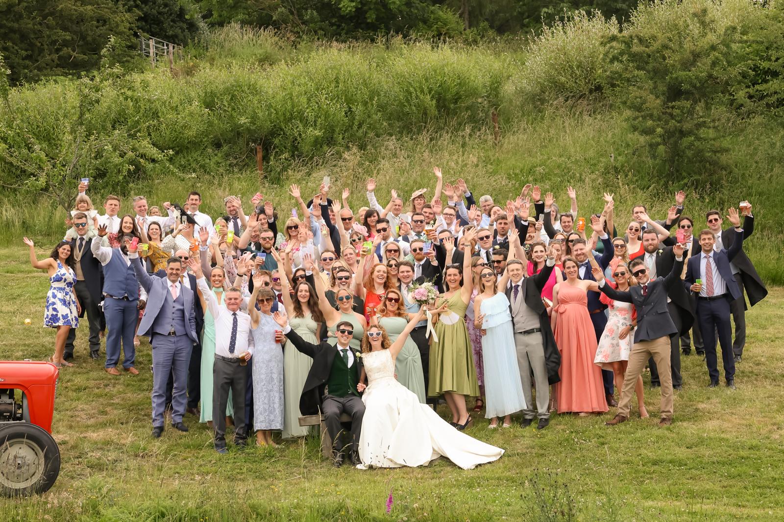 Whole wedding party cheering together in a countryside field beside a vintage tractor
